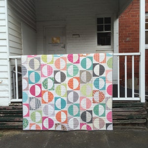 Op de afbeelding: Een kleurrijk quilt met een geometrisch patroon van cirkels en halve cirkels in tinten roze, blauw, groen, oranje en grijs. De quilt hangt aan een wit verandahek.