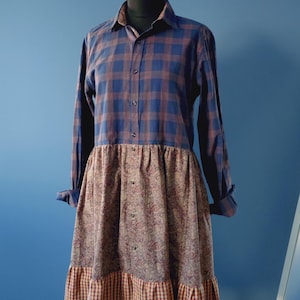 Op de afbeelding: Een unieke jurk met een blauw-bruin geruit overhemd lijfje en een rok met paisley print. De rok heeft een gefranjerde zoom met een bruin-oranje ruitpatroon. De jurk wordt op een mannequin gepresenteerd.