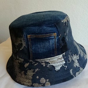 Op de afbeelding: Een blauwe denim bucket hat met een zilver en zwart patroon. De hoed heeft een denim zakje aan de voorkant.