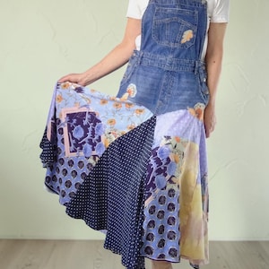 May include: A blue denim overall dress featuring a patchwork skirt made of various fabrics. The skirt includes panels of dark blue fabric with white polka dots, light blue floral patterns, and purple sections with botanical prints.