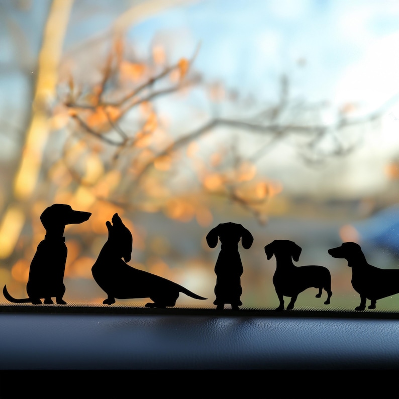 Puede incluir: Cinco calcoman&iacute;as de perro salchicha en silueta negra en la ventana de un coche. Las calcoman&iacute;as est&aacute;n dispuestas en fila, con los perros mirando hacia adelante. Las calcoman&iacute;as son una opci&oacute;n popular para los propietarios de coches que quieren mostrar su amor por sus perros.