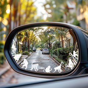 May include: A car side mirror reflecting a street scene with trees and parked cars. White monkey-shaped decals are applied to the mirror's surface, adding a playful touch. The overall image has a sunny, outdoor feel.