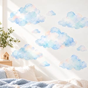 May include: A set of wall decals featuring multiple watercolor-style clouds in shades of light blue, soft white, and pale pink. The clouds vary in size and shape, creating a scattered arrangement across the wall surface above a bed.