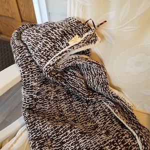 May include: A brown and white knitted throw blanket with a white crocheted border. The blanket is folded and draped over a white chair.