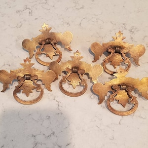 May include: Five antique brass drawer pulls with ornate designs. Each pull has a ring handle and a decorative backplate.