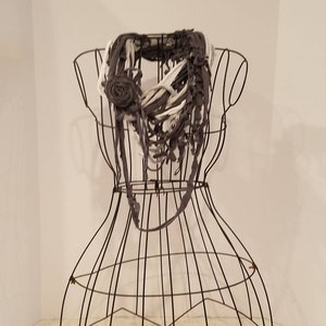 May include: A handmade gray, white, and black scarf with a rose accent, displayed on a wire dress form. The scarf is made of fabric strips and has a fringed design. The rose is made of the same fabric as the scarf.