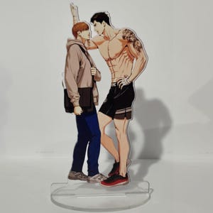 May include: A clear acrylic stand with a vibrant illustration of two male figures. One is shirtless with a tattoo, the other in a hoodie and carrying a bag. The stand has a circular base, suitable for display.