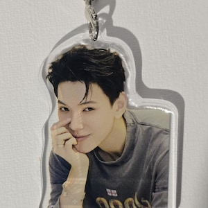 May include: A clear acrylic keychain featuring a smiling person with dark hair, wearing a gray shirt. The keychain has a silver-colored clasp and a split ring for attaching to keys or bags. The shirt has a design at the bottom.