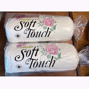 May include: Two cylindrical rolls of white Poly-Fil Soft Touch batting, each encased in clear plastic packaging. The labels feature black cursive text reading Soft Touch, a pink rose graphic with green leaves, and a star logo stating Crafted With Pride In U.S.A.