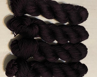 May include: Four skeins of dark purple yarn arranged horizontally. Each skein is tightly twisted into a long, cylindrical shape with a visible ply structure. The yarn has a matte finish and a consistent deep violet color throughout all four units.