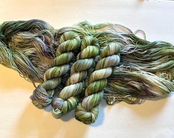 May include: Three skeins of hand-dyed yarn are arranged over a large, loose hank of the same fiber. The yarn features a variegated color palette of sage green, olive, cream, and subtle hints of soft purple and grey throughout the strands.