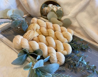 May include: A close-up of four skeins of soft, pale yellow yarn arranged on a wooden tray with green foliage.