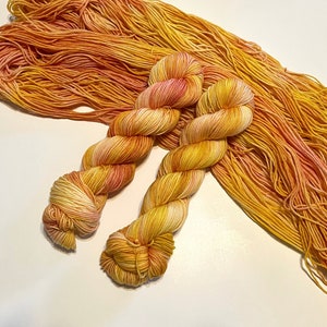 May include: Two skeins of hand-dyed yarn in shades of yellow, orange, and pink. The yarn is a fingering weight and has a subtle gradient.