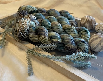 May include: Several skeins of hand-dyed yarn featuring a variegated color palette of deep forest green, charcoal gray, and muted tan. The yarn is arranged in a rectangular wooden tray alongside two decorative spheres and a sprig of faux greenery.