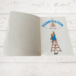 Pode incluir: Um livro aberto intitulado "The Paddington Treasury" com uma imagem de Paddington Bear em uma escada pintando uma parede e uma imagem de Paddington Bear na cama na p&aacute;gina oposta.