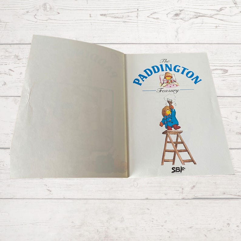 Pode incluir: Um livro aberto intitulado "The Paddington Treasury" com uma imagem de Paddington Bear em uma escada pintando uma parede e uma imagem de Paddington Bear na cama na p&aacute;gina oposta.