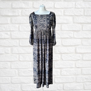 May include: A long, dark blue dress with a square neckline and long sleeves. The dress features a repeating floral and geometric pattern in cream and dark blue. The dress is displayed on a mannequin against a white brick wall.