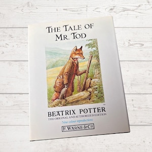 May include: A book titled The Tale of Mr. Tod by Beatrix Potter, featuring an illustration of a fox wearing a brown coat and holding a walking stick. The cover includes the text The Original and Authorized Edition and F. Warne & Co.