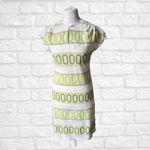 May include: A vintage dress displayed on a mannequin. The dress has a white base with green oval patterns and horizontal stripes. It has short sleeves and a keyhole neckline with a button. The dress is a retro style.