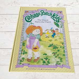 May include: A yellow book with a colourful illustration of a Cabbage Patch Kid doll on the cover. The title of the book is "Cabbage Patch Kids" and the subtitle is "The Shyest 'Kid in the 'Patch". The book is by Kathleen N. Daly and illustrated by Lynn Lace.