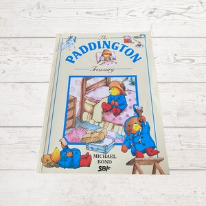 Pode incluir: A capa do livro "The Paddington Treasury" apresenta uma ilustra&ccedil;&atilde;o colorida de Paddington Bear com um casaco azul e botas vermelhas. O t&iacute;tulo do livro &eacute; "The Paddington Treasury" e o autor &eacute; Michael Bond.