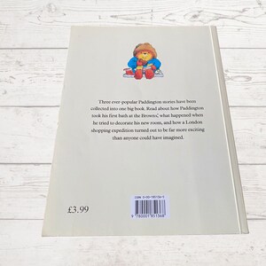 Pode incluir: Uma capa de livro com Paddington Bear vestindo um casaco vermelho e um chap&eacute;u azul. O t&iacute;tulo do livro &eacute; "Paddington" e o texto na contracapa descreve o livro como uma cole&ccedil;&atilde;o de tr&ecirc;s hist&oacute;rias de Paddington. O n&uacute;mero ISBN &eacute; 0-00-185136-5.