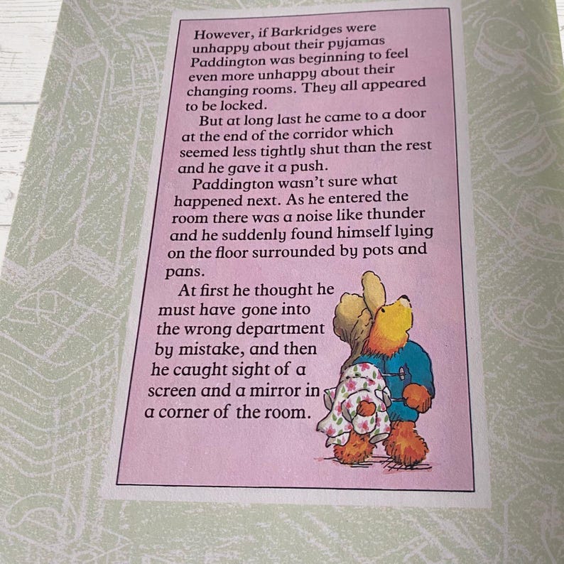 Pode incluir: Uma p&aacute;gina ilustrada de um livro com o personagem Paddington Bear. O texto diz: "No entanto, se os Barkridges estavam infelizes com seus pijamas, Paddington estava come&ccedil;ando a se sentir ainda mais infeliz com seus vesti&aacute;rios. Todos pareciam estar trancados. Mas finalmente ele chegou a uma porta no final do corredor que parecia menos fechada que as outras e a empurrou. Paddington n&atilde;o tinha certeza do que aconteceu em seguida. Ao entrar na sala, houve um barulho como um trov&atilde;o e ele de repente se viu deitado no ch&atilde;o cercado por panelas e frigideiras. No in&iacute;cio, ele pensou que devia ter entrado no departamento errado por engano, e ent&atilde;o viu uma tela e um espelho em um canto da sala."