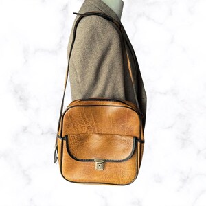 May include: A vintage brown leather crossbody bag with black trim and a front flap. The bag has a rectangular shape with a shoulder strap and a metal clasp closure. The bag is worn over a brown tweed jacket.