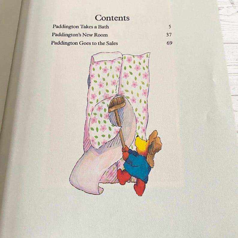 Pode incluir: Um urso castanho com um chap&eacute;u vermelho e uma jaqueta azul est&aacute; segurando uma vassoura e puxando um papel de parede estampado em rosa e verde. O texto na p&aacute;gina diz "Contents", "Paddington Takes a Bath", "5", "Paddington's New Room", "37", "Paddington Goes to the Sales", "69".