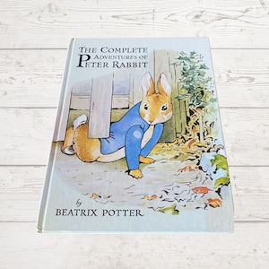 以下が含まれることがあります： ピーターラビットの冒険全集の表紙。青い服を着たウサギが木の柱の後ろから顔を出している。表紙は薄い青色で、背景は白。タイトルは黒文字で書かれている。