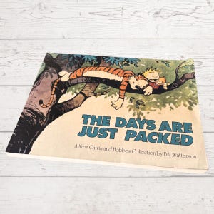 May include: A book titled "THE DAYS ARE JUST PACKED" with a cartoon illustration of Calvin and Hobbes. The cover art depicts the tiger Hobbes and Calvin asleep on a tree branch. The book is a collection by Bill Watterson.