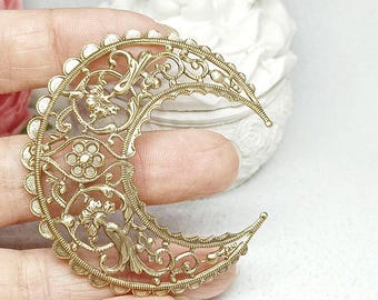May include: A gold-colored filigree crescent moon pendant with intricate floral and scrollwork details. The pendant has a scalloped edge and is held in a hand against a blurred background.