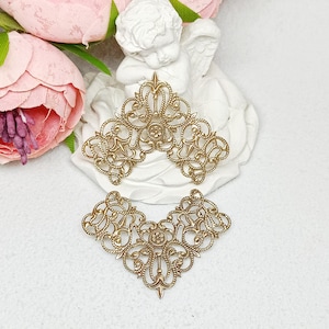 May include: Two ornate, gold-colored filigree embellishments. One is a half-circle shape, the other is a V-shape. A white cherub statue and pink flowers are in the background. These decorative pieces are likely for crafting or jewelry making.