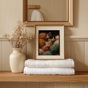 May include: A framed print featuring three kittens nestled among colorful yarn balls. The artwork is displayed on a wooden shelf, alongside a vase of dried flowers and two folded white towels. A gold-framed mirror hangs above.