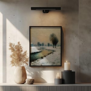 Könnte beinhalten: Gerahmtes Landschaftsgemälde einer verschneiten Flusslandschaft mit Bäumen. Über dem Kunstwerk ist eine schwarze Leuchte angebracht. Eine Vase mit getrockneten Pflanzen, Kerzen und eine dekorative Schale stehen auf einem Regal.
