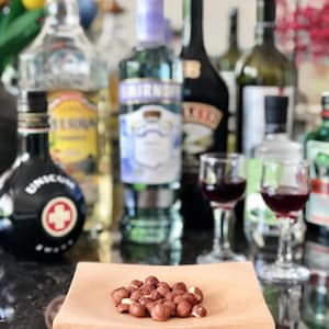 May include: A square, light-colored wooden bowl containing a small pile of raw, brown hazelnuts. The bowl is positioned in the foreground, while several glass bottles of various alcoholic beverages and two small glasses of red liquid are blurred.