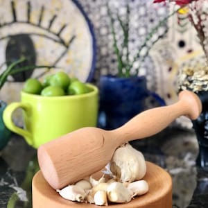 May include: A light-colored wooden mortar and pestle set featuring a circular base and a smooth, rounded handle. Several white garlic cloves are placed in the center of the wooden base, partially covered by the pestle, showcasing a natural wood grain.