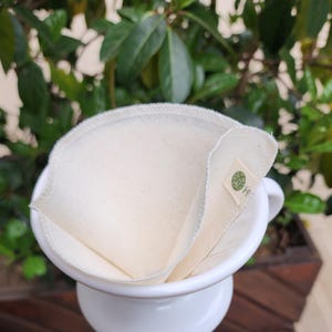 Op de afbeelding: Een kegelvormig, gebroken wit stoffen koffiefilter met een gestikte rand, voorzien van een klein rechthoekig label met een groen cirkelvormig logo. Het filter zit in een witte keramische koffiefilterhouder op een glazen karaf.