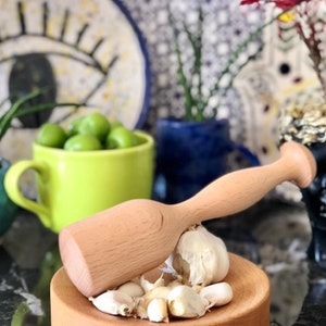 Op de afbeelding: Een houten knoflookpers met een ronde basis en een lange handgreep. De pers is gemaakt van lichtbruin hout en staat op een zwart oppervlak met een stapel knoflookteentjes.