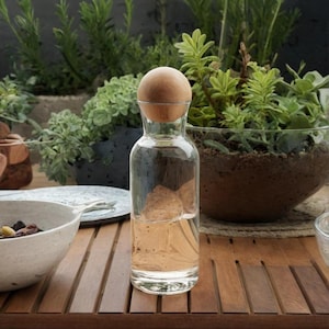 May include: A clear glass bottle with a wooden ball stopper. The bottle is filled with water and is sitting on a wooden table.