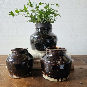 May include: Three brown glazed ceramic jars with a dark brown glaze. The jars are of varying sizes and are arranged on a wooden surface. A green plant is in the largest jar.