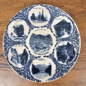 May include: A round, off-white ceramic plate with a blue and white design. The plate features illustrations of landmarks around Pikes Peak, including Balanced Rock, Cathedral Spires, and Seven Falls. The text "Pikes Peak & Vicinity" is also visible.