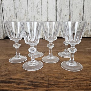 May include: Six clear, stemmed glasses with a cut-glass design. The glasses have a flared bowl, a faceted stem, and a round base. The glasses are arranged on a wooden surface.