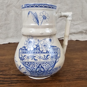 May include: Cream-colored ceramic pitcher with blue floral and geometric patterns. The pitcher features a handle and a spout. The design includes flowers, leaves, and decorative borders. The pitcher is likely vintage or antique.