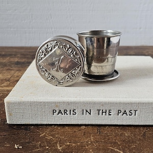 Può includere: Una tazza pieghevole color argento e la sua custodia abbinata. La custodia è rotonda con un bordo ornato e un centro a forma di diamante con la lettera "K". La tazza è accanto alla custodia su un libro intitolato "PARIS IN THE PAST".