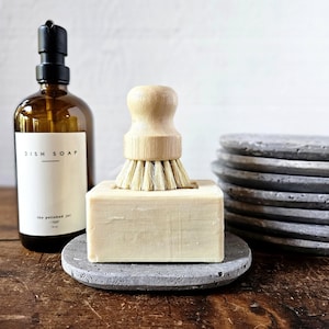 May include: A brown glass bottle labeled "DISH SOAP" with a black pump dispenser. A wooden handled dish brush sits atop a rectangular soap bar on a gray stone soap dish. A stack of gray stone coasters is in the background.