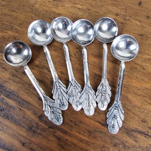 May include: A set of six silver-colored spoons with round bowls and leaf-shaped handles. The spoons are arranged on a wooden surface, showcasing their reflective surfaces and intricate design. Ideal for serving or display.