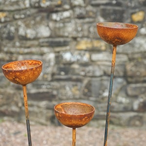 May include: Three rusty metal planters with a rounded bowl shape and a long stem. The planters are arranged in a triangular formation against a stone wall background.