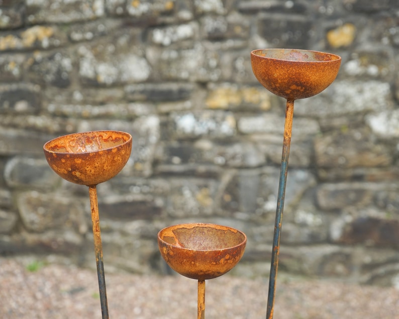 May include: Three rusty metal planters with a rounded bowl shape and a long stem. The planters are arranged in a triangular formation against a stone wall background.