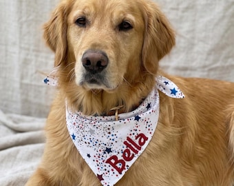 May include: A golden retriever wearing a white bandana with a red, white, and blue star pattern. The name "Bella" is printed in red on the bandana. The dog is lying down and looking at the camera.
